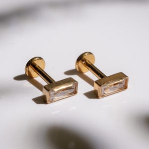 May include: Two gold-colored stud earrings with rectangular, clear gemstones. The earrings have a flat, round backing and a post that connects to the rectangular gemstone setting. The earrings are displayed on a white surface.