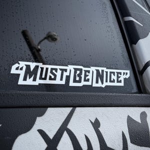 May include: A white sticker on a car window displaying the phrase "MUST BE NICE" in bold, capitalised letters. The sticker is on a black surface with a black and white graphic design.