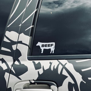 Puede incluir: Una camioneta de camuflaje blanco y negro con una pegatina blanca de una vaca con la palabra "BEEF" en ella.