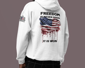 Puede incluir: Sudadera con capucha blanca con un estampado gráfico de una bandera estadounidense en colores rojo, blanco y azul. El texto "FREEDOM IS NEVER GIVEN. IT IS WON" está impreso en la sudadera.