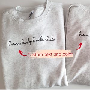 May include: Two grey sweatshirts with the text "homebody book club" in black cursive lettering. The text is customisable.
