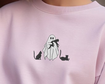 May include: A pink sweatshirt with a white ghost holding a black cat. Two black cats are standing on either side of the ghost.