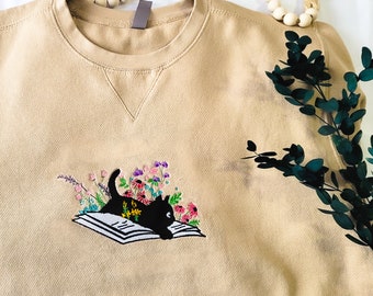 May include: A beige sweatshirt with an embroidered design of a black cat reading a book surrounded by colorful flowers.