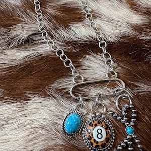 May include: A silver-toned chain necklace featuring an oval connector link with three hanging charms: an oval turquoise stone in a twisted rope setting, a leopard-print billiard ball charm with the number eight, and a beaded cross with a turquoise center.