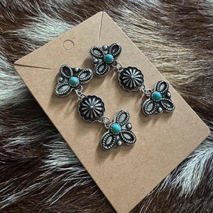 May include: Silver-toned earrings with turquoise-colored stones and a floral design. The earrings are displayed on a brown card against a brown and white patterned background. The earrings have a dangling design.