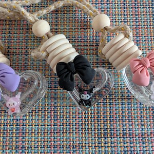 May include: Three heart-shaped car air fresheners with decorative bows and character figurines. Each freshener has a clear heart-shaped container, a bow, and a small character figurine. The bows are purple, black, and pink. The fresheners hang from a beige rope.