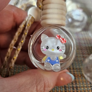 Könnte beinhalten: Transparenter Auto-Lufterfrischer aus Glas mit einer Cartoon-Katzenfigur im Inneren. Die Katze hat eine rote Schleife, blaue Latzhosen und gelbe Akzente. Der Lufterfrischer hat einen Holzdeckel und eine geflochtene Kordel zum Aufhängen.