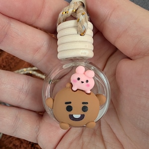 May include: A clear glass car air freshener with a wooden top and a decorative cartoon character. The character is brown with a pink bunny on top. The air freshener has a braided gold and beige string for hanging.