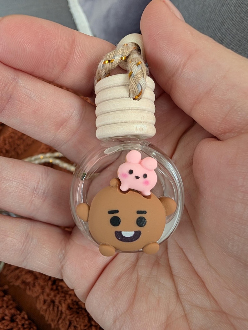May include: A clear glass car air freshener with a wooden top and a decorative cartoon character. The character is brown with a pink bunny on top. The air freshener has a braided gold and beige string for hanging.