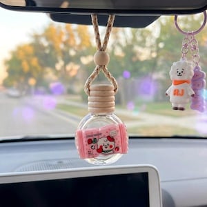 Può includere: Un flacone diffusore per auto in vetro trasparente con tappo in legno e cordino intrecciato, caratterizzato da un'etichetta rosa con un personaggio e il testo RJ. Accanto pende un portachiavi viola con un personaggio bianco e la scritta ARMY.