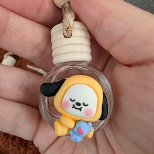 May include: A clear glass bottle with a wooden cap and a decorative charm inside. The charm is a yellow cartoon character with black ears, a white face, and a blue stuffed animal. The bottle is suspended by a braided cord.