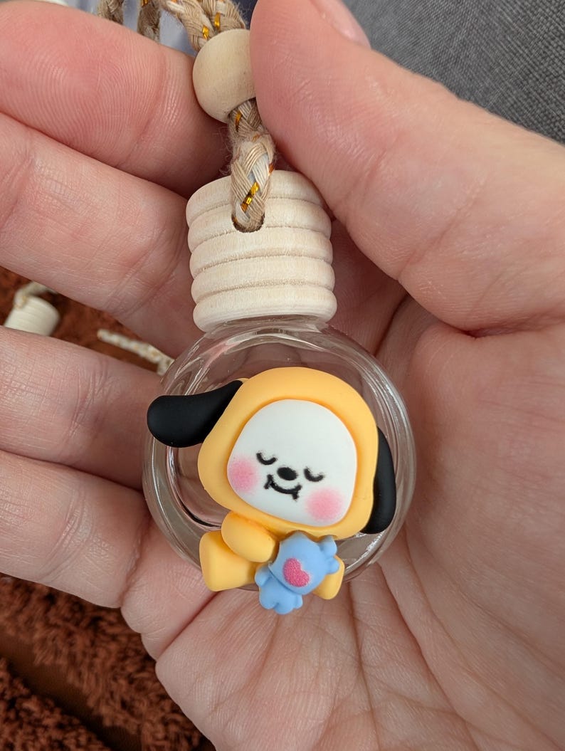 May include: A clear glass bottle with a wooden cap and a decorative charm inside. The charm is a yellow cartoon character with black ears, a white face, and a blue stuffed animal. The bottle is suspended by a braided cord.