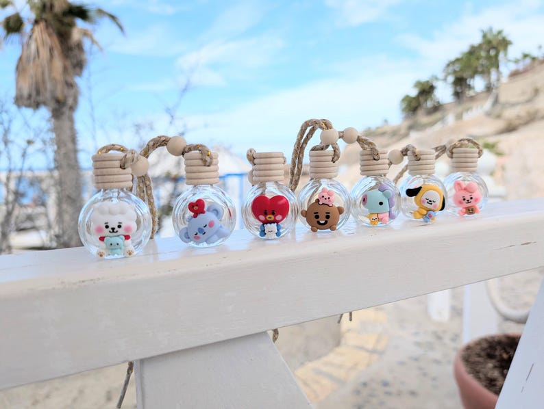 May include: A row of seven clear glass aromatherapy bottles with cartoon character designs. Each bottle has a wooden cap, a wooden bead, and a woven cord. The background shows a beach scene with a blue sky and palm trees.