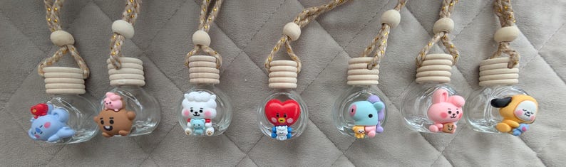 May include: A collection of seven clear glass car air fresheners, each with a unique cartoon character figurine inside. The figurines are in various colors, including blue, brown, red, and yellow, and are suspended from a wooden top with a decorative string.