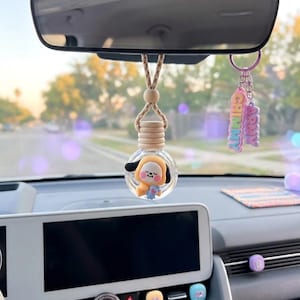 Op de afbeelding: Auto-interieur met een hangende luchtverfrisser in een glazen fles, met een stripfiguur. Een sleutelhanger met "CHIMMY ARMY" hangt ernaast. Het dashboard bevat een knop met het opschrift "MAGIC SHOP".