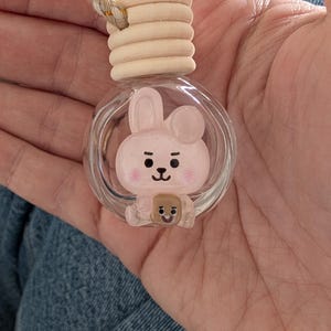 May include: A clear glass car air freshener with a cartoon bunny design inside. The bunny is pink with black details and a small brown bear. The air freshener has a wooden top and a beige rope for hanging.