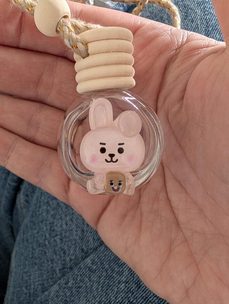 May include: A clear glass car air freshener with a cartoon bunny design inside. The bunny is pink with black details and a small brown bear. The air freshener has a wooden top and a beige rope for hanging.
