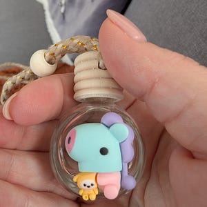 May include: A clear glass air freshener bottle with a light blue, pink, and purple cartoon character inside. The bottle has a wooden top and a beige and gold rope for hanging.