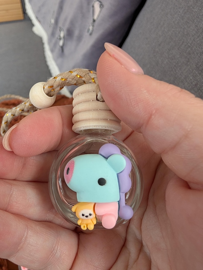 May include: A clear glass air freshener bottle with a light blue, pink, and purple cartoon character inside. The bottle has a wooden top and a beige and gold rope for hanging.