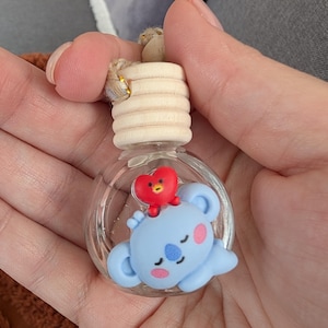 May include: A clear glass aromatherapy bottle with a light wood cap and a small, light blue koala figure with a red heart on top. The koala has pink cheeks and a blue nose. The bottle is suspended by a natural fiber cord.