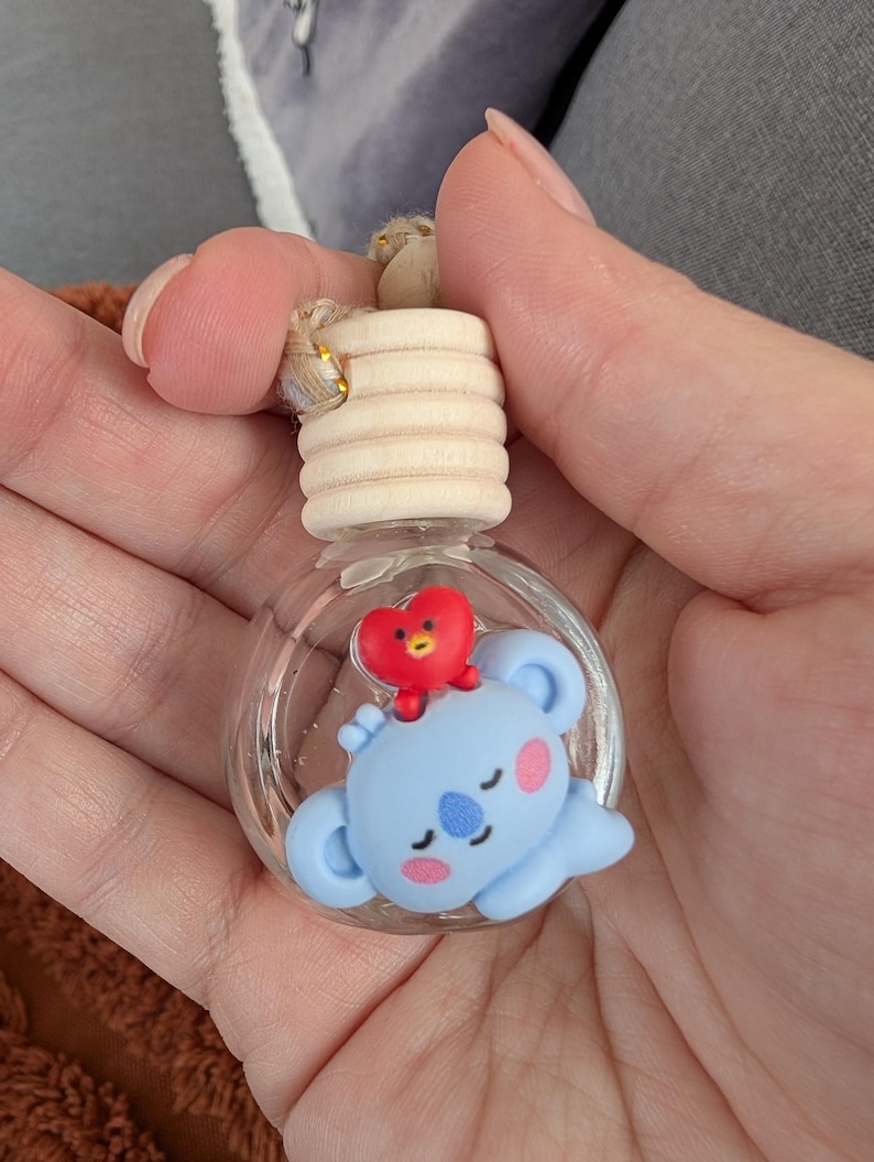 May include: A clear glass aromatherapy bottle with a light wood cap and a small, light blue koala figure with a red heart on top. The koala has pink cheeks and a blue nose. The bottle is suspended by a natural fiber cord.