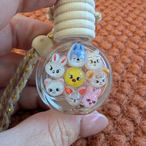 Puede incluir: Un ambientador de coche de cristal transparente con una parte superior de madera y un cordón decorativo. El ambientador contiene varias caras de animales de dibujos animados pequeños y coloridos. Las caras incluyen un conejo, un oso, un perro y un pollito.