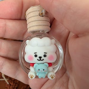 May include: A clear glass bottle with a wooden cap, featuring a white cartoon character with pink cheeks and a small blue bear. The character has a red collar and paw prints. The bottle is designed for aromatherapy.