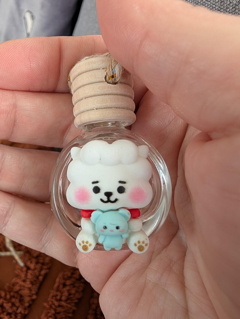 May include: A clear glass bottle with a wooden cap, featuring a white cartoon character with pink cheeks and a small blue bear. The character has a red collar and paw prints. The bottle is designed for aromatherapy.