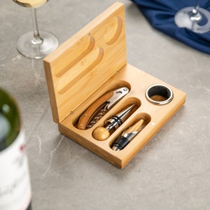 May include: A wooden box with a set of wine tools, including a corkscrew, a bottle stopper, and a foil cutter. The tools are arranged in the box, which is open and has a light wood finish.
