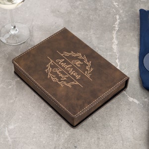 May include: A brown leatherette box with the engraved text "The Anderson Family" framed by a decorative wreath. The box is rectangular with a stitched border. Wine glasses and a bottle of wine are in the background.