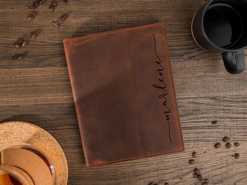 May include: A brown leather journal with the name "marlene" in a cursive font on the right side. The journal is on a wooden surface with coffee beans and a black mug.