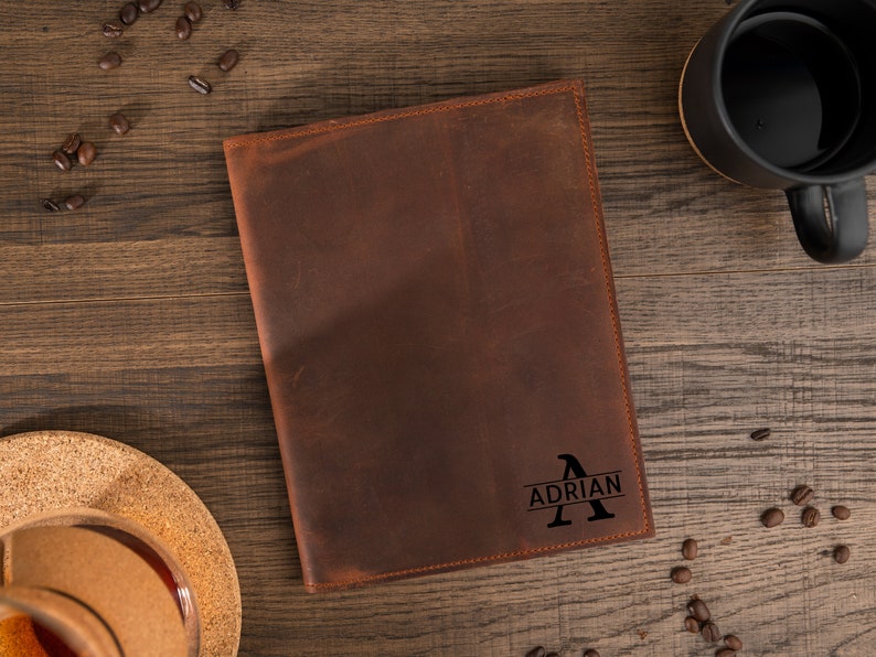May include: A brown leather journal with the name "Adrian" and the letter "A" engraved on the cover. The journal is on a wooden surface, with coffee beans and a black mug in the background.
