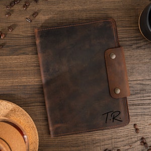 May include: A brown leather journal with a snap closure, featuring the initials "T.R." in the lower right corner. The journal is set on a wooden surface with coffee beans and a black mug. The leather has a distressed look.