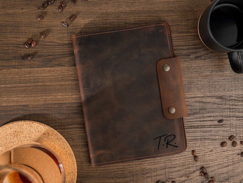 May include: A brown leather journal with a snap closure, featuring the initials "T.R." in the lower right corner. The journal is set on a wooden surface with coffee beans and a black mug. The leather has a distressed look.