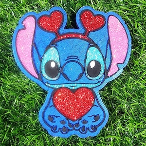 May include: Blue and pink glittery character holding a red glittery heart. The character has large eyes and two red hearts on its head.