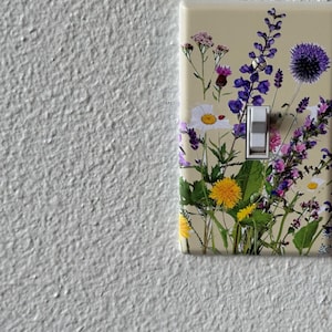 May include: A light switch cover with a floral design featuring purple, white, and yellow flowers on a cream background.