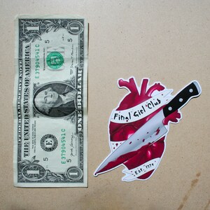 May include: A red heart with a knife through it. The heart has a white banner that says "Final Girl Club" and "Est. 1979".