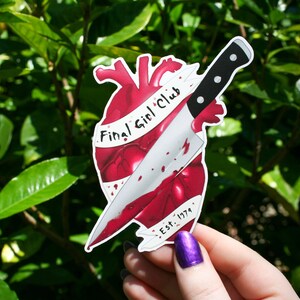May include: A red heart with a bloody knife through it. The knife has a black handle and a silver blade. The heart has a white banner wrapped around it that says "Final Girl Club Est. 1979".