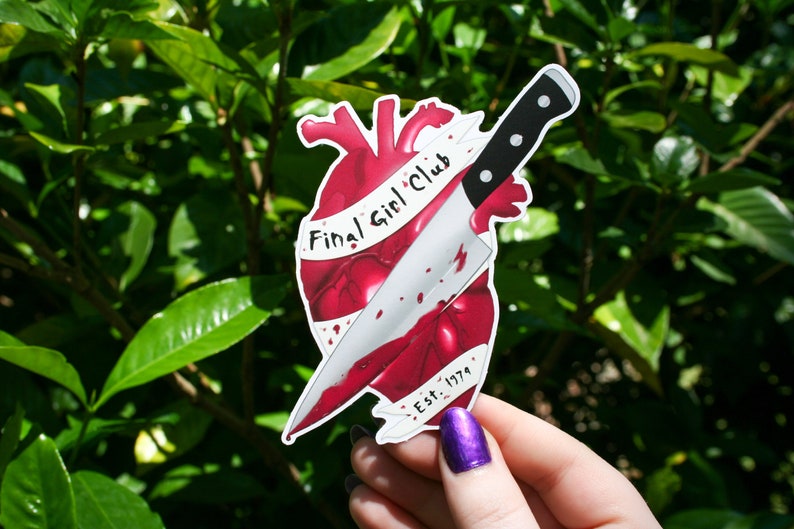 May include: A red heart with a bloody knife through it. The knife has a black handle and a silver blade. The heart has a white banner wrapped around it that says "Final Girl Club Est. 1979".