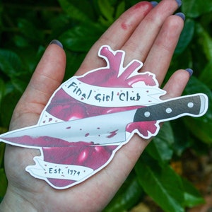 May include: A white sticker featuring a red heart pierced by a bloody knife. The heart has a banner that reads "Final Girl Club Est. 1974".