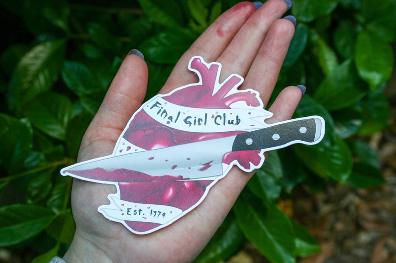 May include: A white sticker featuring a red heart pierced by a bloody knife. The heart has a banner that reads "Final Girl Club Est. 1974".