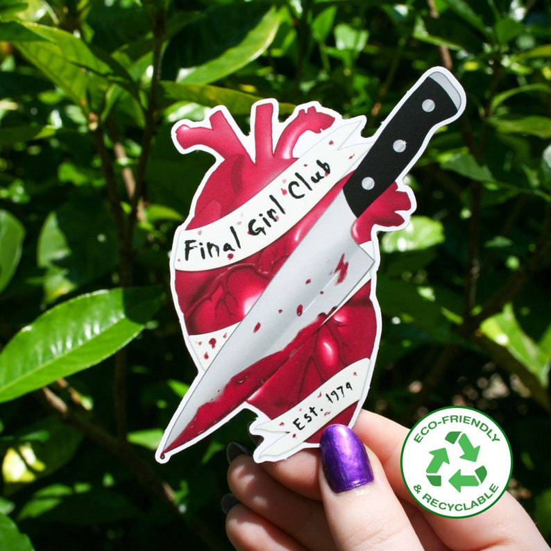 May include: A red heart with a knife sticking through it. The knife has a black handle and a silver blade. The heart has a white ribbon wrapped around it that says "Final Girl Club Est. 1979".