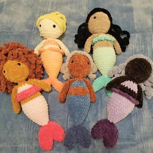 May include: A collection of six handmade crochet mermaid dolls. Each doll features a unique color scheme, including yellow, brown, black, and orange hair, with matching or contrasting tails in shades of pink, blue, and purple. The dolls have detailed facial features.