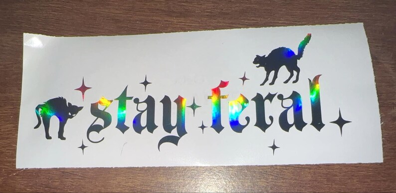 May include: A white sticker with a black silhouette of two cats and the text "stay feral" in a gothic font. The sticker has a rainbow iridescent finish.