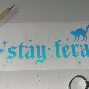 May include: A white vinyl decal with a blue to teal gradient. The decal features the text "stay feral" in a gothic font with two black cat silhouettes. The cat silhouettes are facing left and right.
