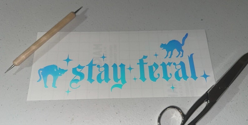 May include: A white vinyl decal with a blue to teal gradient. The decal features the text "stay feral" in a gothic font with two black cat silhouettes. The cat silhouettes are facing left and right.