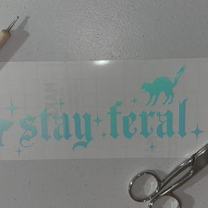 May include: A white vinyl sticker with a teal blue design that says "stay feral" in a gothic font. There are two black cat silhouettes on the sticker. The sticker is on a white surface with a wooden tool and silver scissors.
