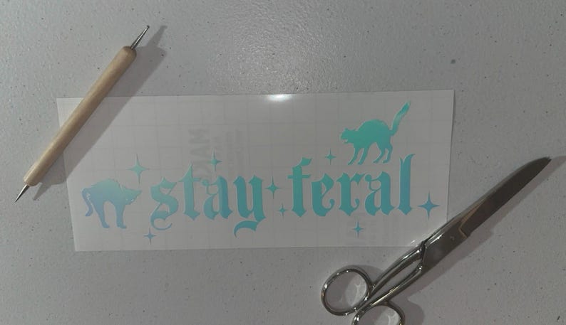 May include: A white vinyl sticker with a teal blue design that says "stay feral" in a gothic font. There are two black cat silhouettes on the sticker. The sticker is on a white surface with a wooden tool and silver scissors.