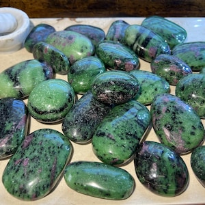 May include: A collection of polished ruby in zoisite stones. The stones are oval-shaped, with a mix of green, black, and pink hues. They are arranged on a light-colored surface, with a white mortar and pestle in the background.