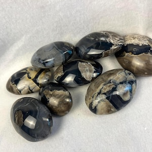 May include: A collection of polished, oval-shaped stones. The stones display a variety of colors, including black, gray, and brown, with intricate patterns and veining. The stones are smooth and reflective, suggesting a natural origin.